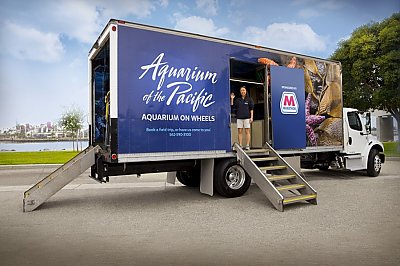 Aquarium on Wheels truck set up for display with open side and back doors with stairs that lead up to tidepool area. Smiling educator greets the camera.